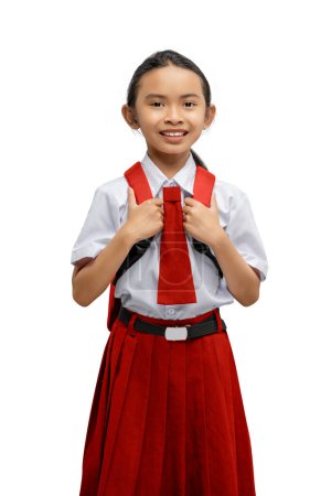 Porträt eines fröhlichen jungen Mädchens in Schuluniform, das sich selbstbewusst an ihre Rucksackgurte klammert, isoliert auf weißem Hintergrund mit Kopierraum