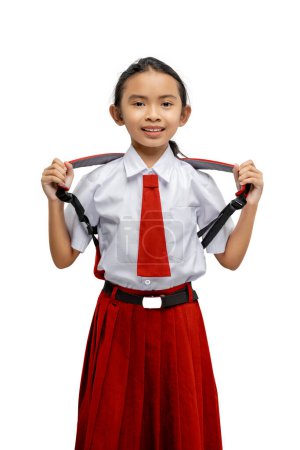 Cheerful young girl stands proudly in her red and white school uniform, playfully pulling on her suspenders, isolated on a white background with a bright smile