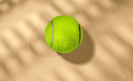Single bright yellow tennis ball lying on a sandy surface with palm tree shadow patterns, highlighting concepts of sport, leisure, summer, and outdoor activities