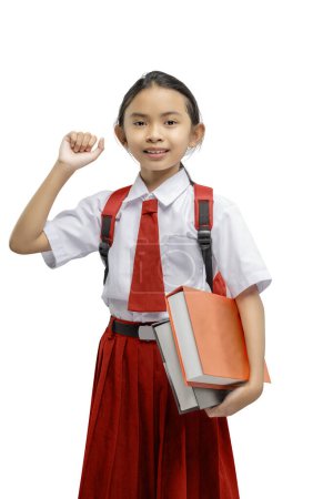 Fröhliches junges Mädchen mit strahlendem Lächeln, in weißem Hemd und roter Rockuniform, hält stolz ihre Bücher in der Hand und zeigt vor weißem Hintergrund Lernfreude