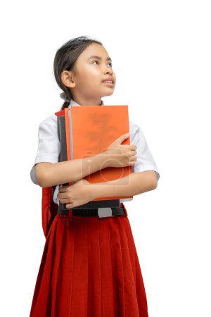 Ein junges Mädchen in Schuluniform mit rotem Rock hält ein orangefarbenes Lehrbuch dicht vor der Brust, während sie mit einem Ausdruck der Kontemplation und Sehnsucht, isoliert auf weißem Hintergrund, nach oben blickt.