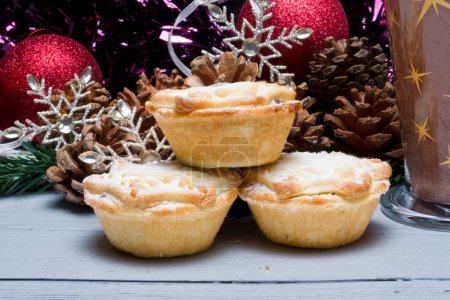 Photo for Three freshly made christmas mince pies surrounded by decorations and glass of hot chocolate off to the side. - Royalty Free Image
