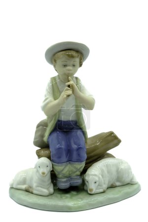Foto de Figura de porcelana antigua de un pastor niño tocando el silbato o las tuberías sentados en un tronco con dos ovejas o corderos a sus pies. - Imagen libre de derechos