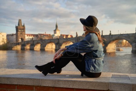 Téléchargez les photos : Stylish young beautiful woman earing black hat in Prague with Charles Bridge on background. Elegant retro lady fine art portrait. - en image libre de droit