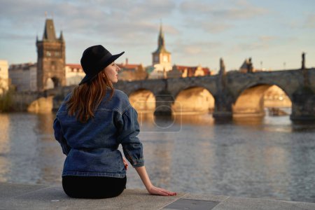 Téléchargez les photos : Stylish young beautiful woman earing black hat in Prague with Charles Bridge on background. Elegant retro lady fine art portrait. - en image libre de droit