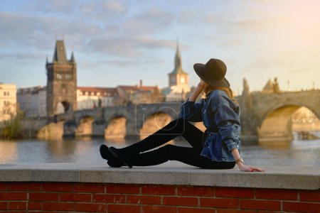 Foto de Elegante joven hermosa mujer con sombrero negro en Praga con el puente de Carlos en el fondo. Elegante retro dama retrato de arte. - Imagen libre de derechos