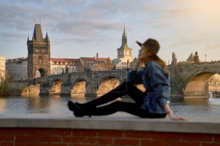 Photo for Stylish young beautiful woman earing black hat in Prague with Charles Bridge on background. Elegant retro lady fine art portrait. - Royalty Free Image