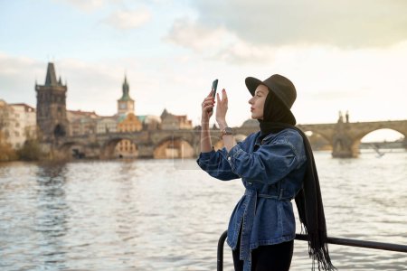 Foto de Elegante joven hermosa mujer con sombrero negro en Praga con el puente de Carlos en el fondo. Elegante retro dama retrato de arte. - Imagen libre de derechos