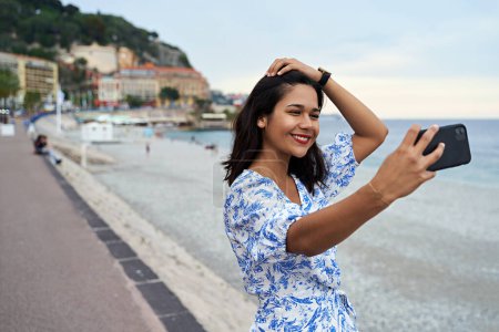 Téléchargez les photos : Belle jeune femme souriante prend selfie avec un smartphone à la Promenade des Anglais à Nice, France - en image libre de droit
