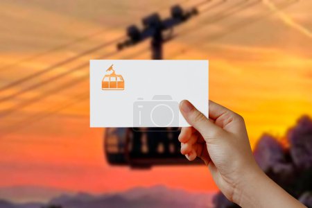 Téléchargez les photos : Man's hand holding cable car symbol paper in sky. Concept of journey, travel, dream, freedom. Hand is holding paper cable car against a rope car with empty space for text. - en image libre de droit