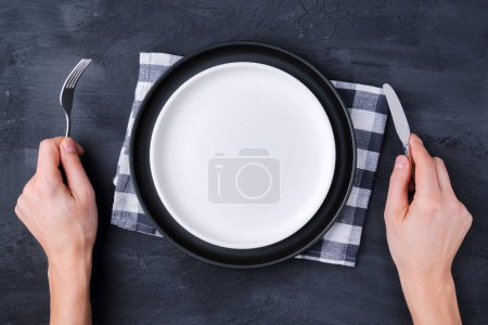 Homme avec une fourchette, un couteau et une assiette vide sur fond noir, vue de dessus