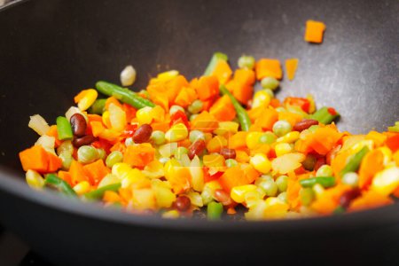 Colorful chopped vegetables is being stirred using a wooden spoon. The vegetables sizzle and release aromatic scents as they cook.