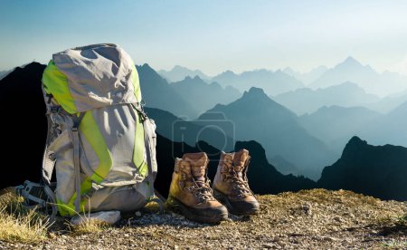 Foto de Hiking equipment with blue moutain silhouettes. Backpack and hiking shoes on top of the mountain. Beautiful view to mountain ranges and fog filled valleys. Tirol, Austria, Allgaeu, Alps. - Imagen libre de derechos