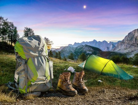 Foto de Illuminated camping tent at night with Backpack and hiking Shoes. High altitude alpine landscape. Shining Moon in dark blue sky with purple clouds. Triglav National Park, Slovenia. Concept for - Imagen libre de derechos