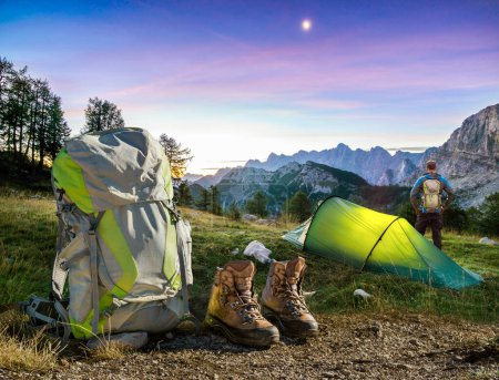 Foto de Illuminated camping tent at night with Backpack and hiking Shoes and a Hiker man. High altitude alpine landscape. Shining Moon in dark blue sky with purple clouds. Triglav National Park, Slovenia - Imagen libre de derechos