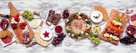 Christmas charcuterie table scene over a white wood banner background. Assortment of cheese and meat appetizers. Christmas tree, wreath and candy cane arrangements.