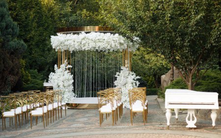 Photo for Luxurious yellow-gold wedding arch decorated with white flowers in nature among the trees. wedding ceremony. white piano on the nature. chairs for guests on the street. - Royalty Free Image
