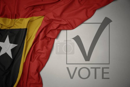 Foto de Ondeando colorida bandera nacional de Timor Oriental sobre un fondo gris con voto de texto. concepto de elección. Ilustración 3D - Imagen libre de derechos