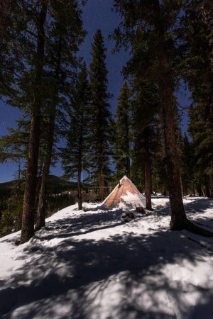 Winter Tent Camping - White Teepee with chimney surrounded  by tall trees 