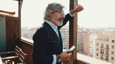Senior disfruta de la vista de pie en el balcón con teléfono móvil en la mano