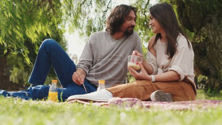 Photo for Happy couple testing snacks while sitting on a blanket in the park - Royalty Free Image