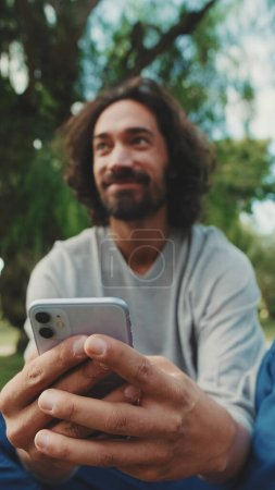 Photo for Young man using mobile phone while sitting on the lawn in the park - Royalty Free Image