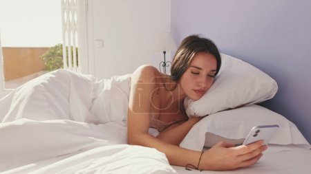 Photo for Happy woman texting on phone after waking up in the morning. Smiling girl having an online conversation, replying tap message while sitting in bed at home - Royalty Free Image