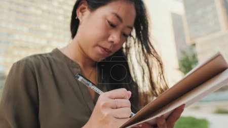 Photo for Young girl looks through notes in notebook while sitting on modern buildings background - Royalty Free Image