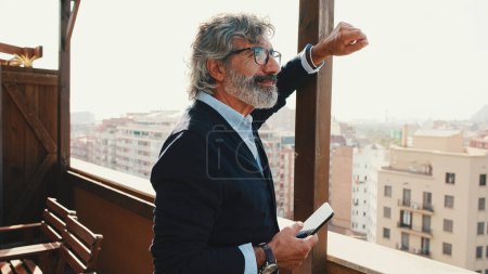 Téléchargez les photos : Senior bénéficie de la vue debout sur le balcon avec téléphone portable à la main - en image libre de droit