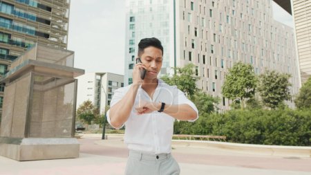 Photo for Young businessman talking on smartphone and looking at watch on modern buildings background - Royalty Free Image