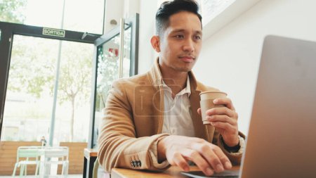 Photo for Young businessman wearing beige suite sits at table with laptop and drinks coffee - Royalty Free Image