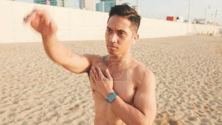 Photo for Guy, the athlete standing on the beach with headphones does a shoulder exercise, makes hand swings while standing on the beach - Royalty Free Image