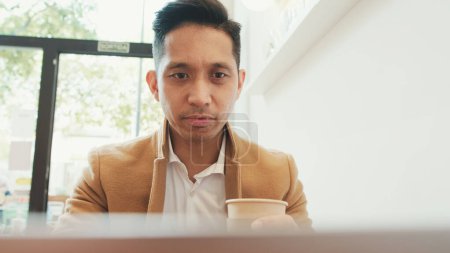Photo for Young businessman wearing beige suite sits at table with laptop and drinks coffee - Royalty Free Image