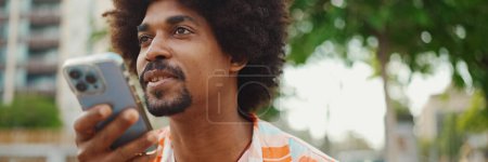 Téléchargez les photos : Portrait en gros plan d'un jeune Afro-Américain portant une chemise assise sur un banc de parc à l'aide de son smartphone. Un homme souriant envoie un message vocal sur son téléphone portable. - en image libre de droit