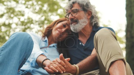 Photo for Happy retired couple sitting in the park holding hands and smiling resting on the grass in the park in autumn - Royalty Free Image