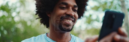 Téléchargez les photos : Gros portrait du jeune Afro-Américain en t-shirt bleu clair assis sur le banc du parc de la ville et utilisant son smartphone. L'homme regarde des photos, des vidéos sur son téléphone portable. concept mode de vie. - en image libre de droit