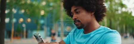 Téléchargez les photos : Gros plan du jeune Afro-Américain en t-shirt bleu assis sur le banc du parc et parlant sur haut-parleur. Un homme souriant envoie un message vocal sur son téléphone portable. concept mode de vie. - en image libre de droit