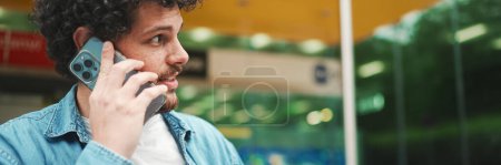 Photo for Young bearded man in denim shirt stands at bus stop waiting for bus and talking on mobile phone on modern city background - Royalty Free Image