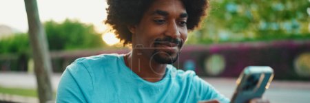 Close-up of young African American man in blue t-shirt sitting on park bench and talking on speakerphone. Smiling man sends voice message on mobile phone. lifestyle concept.