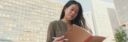 Photo for Young girl looks through notes in notebook while sitting on modern buildings background, Panorama - Royalty Free Image
