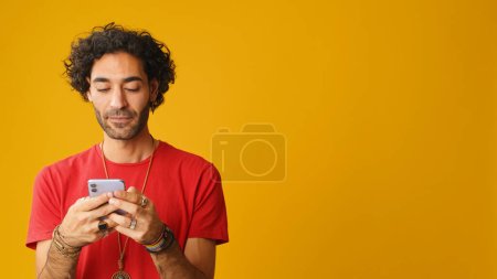 Lächelnder Mann mit lockigem Haar, in rotem T-Shirt gekleidet, benutzt Handy, tippt Nachricht isoliert auf gelbem Hintergrund im Studio