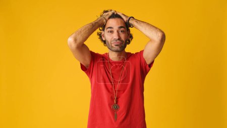 Aufgebrachter unzufriedener Mann mit lockigem Haar, rotes T-Shirt, Blick in die Kamera auf gelbem Hintergrund im Studio