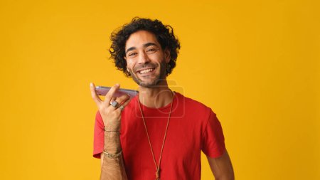 Mann mit lockigem Haar trägt rotes T-Shirt, spricht über Lautsprecher, sendet Sprachnachricht Handy isoliert auf gelbem Hintergrund im Studio