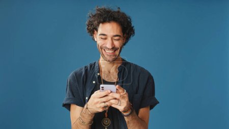 Lächelnder Mann mit lockigem Haar, blaues Hemd, Mobiltelefon, scrollende Anwendung isoliert auf blauem Hintergrund im Studio