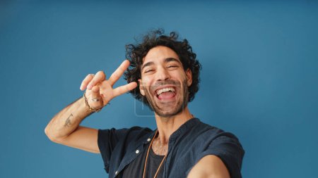 Lächelnder Mann mit lockigem Haar, in blauem Hemd, schaut in die Kamera, die ein Selfie macht, das ein Zeichen des Sieges und der Freiheit auf blauem Hintergrund im Studio zeigt.