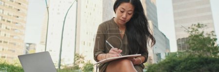 Photo for Young girl uses laptop pc makes notes in notebook from outside on modern buildings background, Panorama - Royalty Free Image