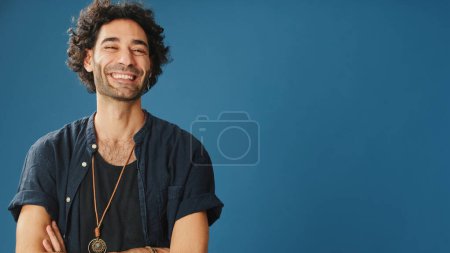 Photo for Man with curly hair, dressed in blue shirt, looks at camera and smiling, isolated on blue background in studio - Royalty Free Image