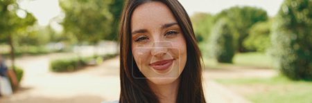 Photo for Close-up of young smiling woman with long brown hair wearing white crop top posing for the camera in the park, Panorama. The girl opens her eyes and smiles at the camera - Royalty Free Image