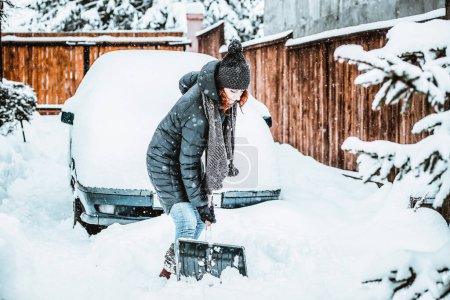 Photo for Woman with shovel cleaning snow around car. Winter shoveling. Removing snow after blizzard - Royalty Free Image
