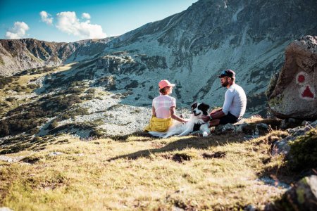Téléchargez les photos : Couple avec chien assis sur des rochers admirant le paysage de montagne - en image libre de droit